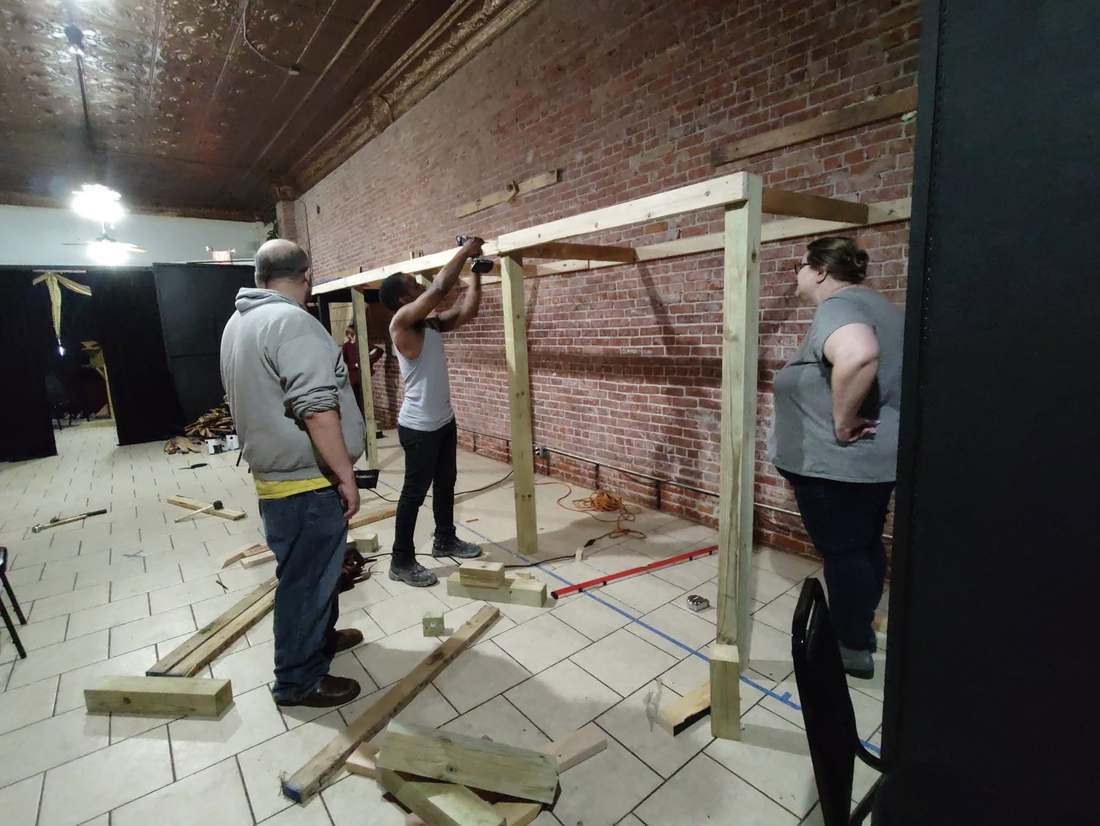 Dream Theatre Co. members building a set in the brick studio — one climbing through a window flat while another works from a ladder on the backdrop
