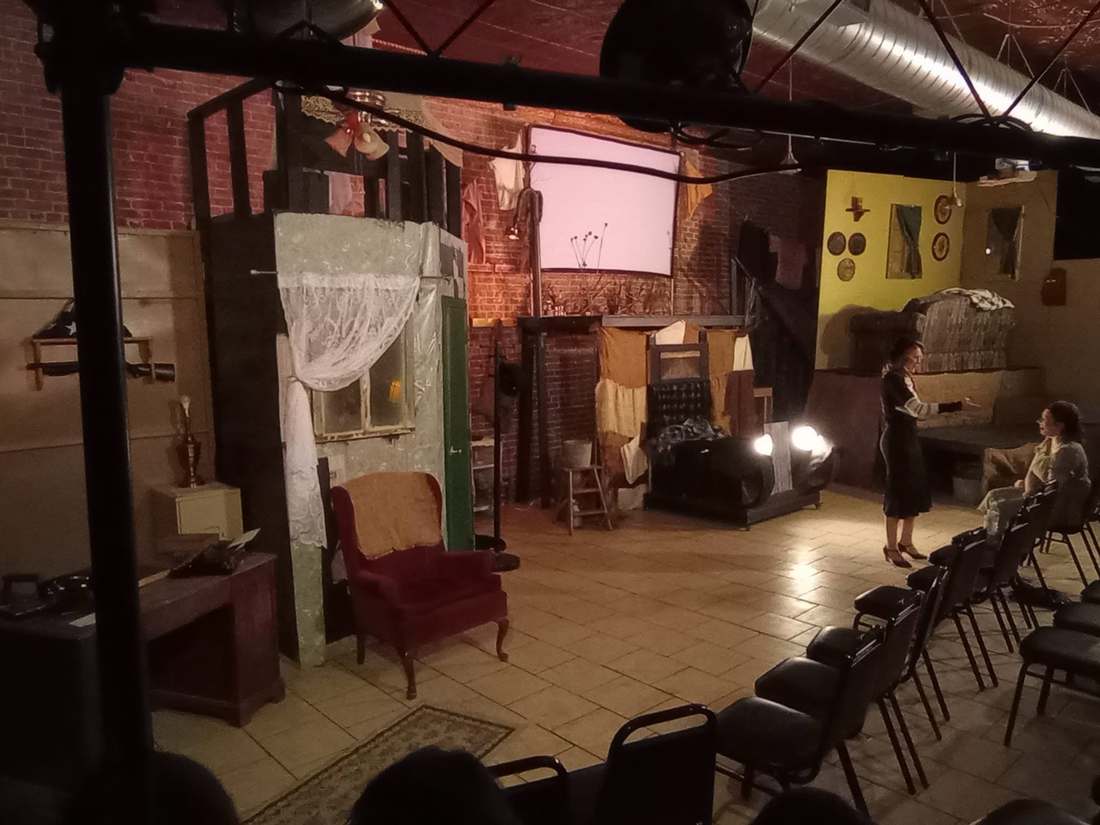 The Dream Theatre Co. studio stage dressed with a set — a red armchair, a lace-curtained window flat, and performers under the lights