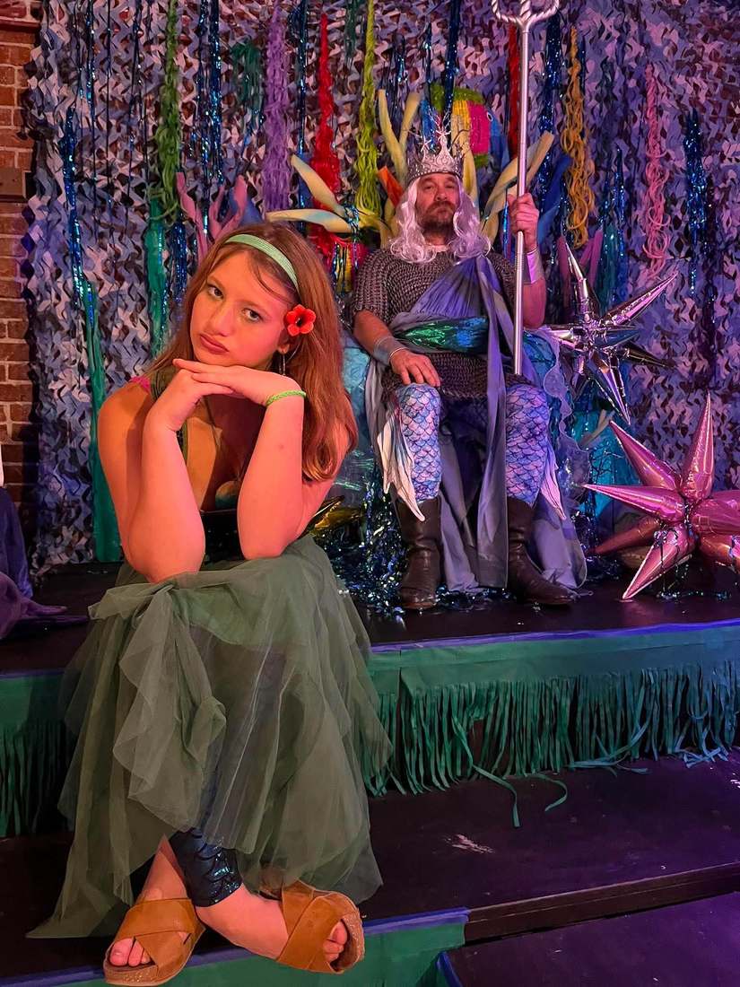 Ariel in a shell top and teal mermaid tail beside King Triton on his throne