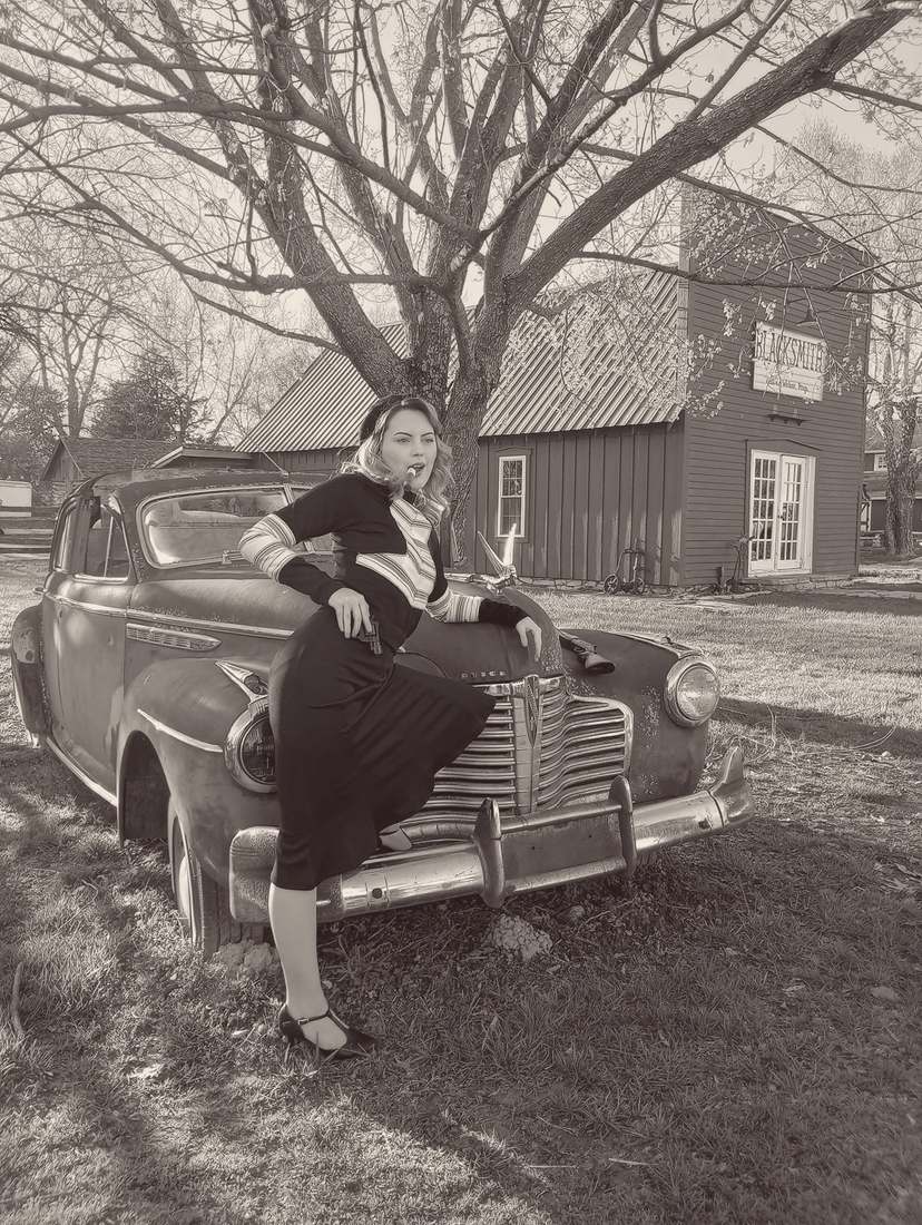 Bonnie posing on the hood of a vintage Buick with a cigar, outside a blacksmith shop (sepia)