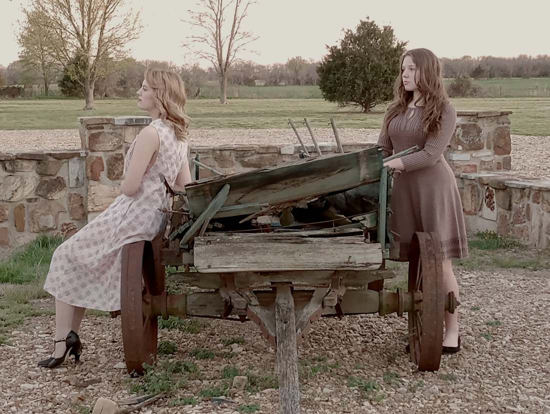 Two cast members in period dresses posing with a weathered farm wagon