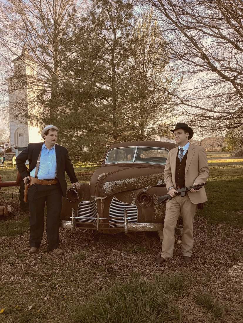 Two cast members in suits standing guard on either side of the rusted vintage car