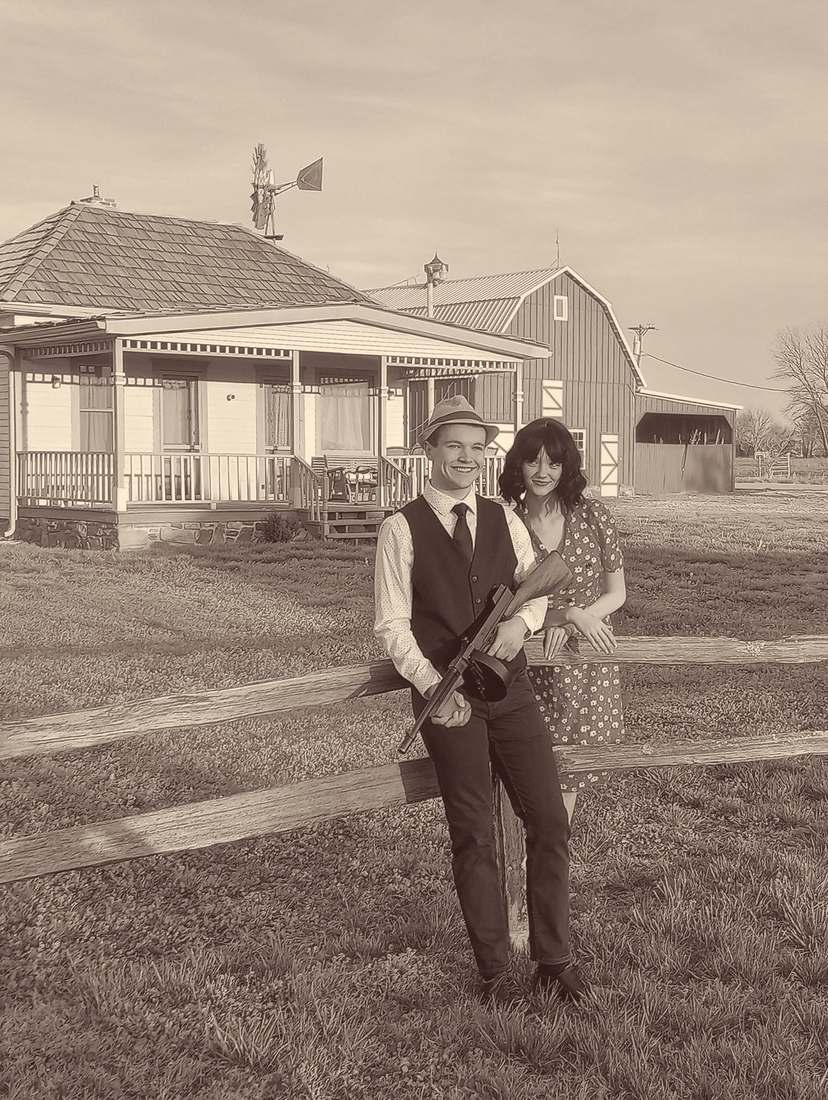 Two cast members smiling at a split-rail fence in front of the farmhouse, one holding a Tommy gun (sepia)