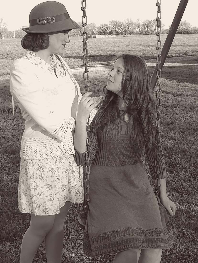 Two cast members talking beside a swing set, one wearing a cloche hat (sepia)