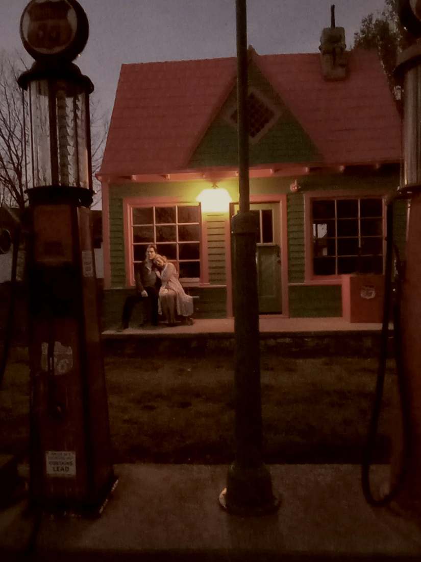 Bonnie resting her head on Clyde's shoulder outside a vintage gas station at night