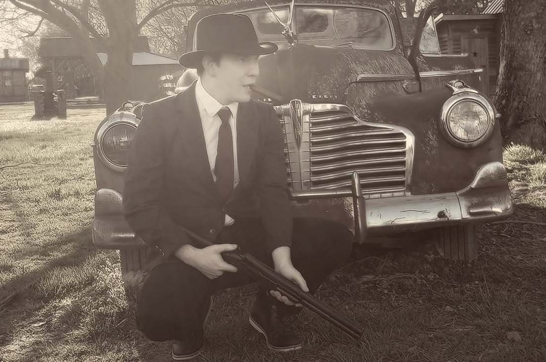 Clyde crouching with a rifle in front of the vintage Buick (sepia)