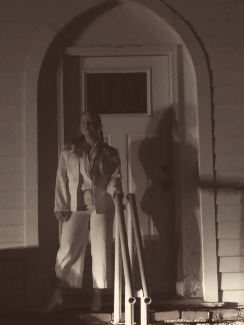 A cast member in a white suit standing in a church doorway at night, her shadow cast on the door (sepia)