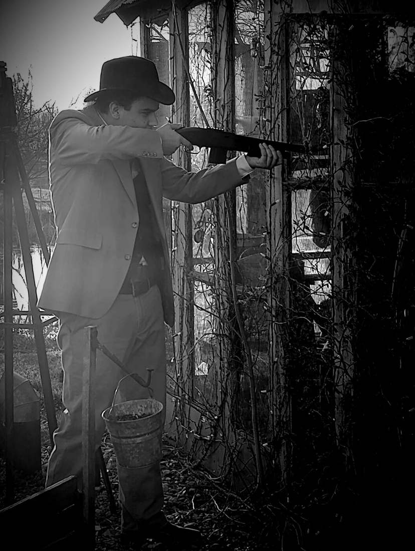 A cast member in a suit and fedora aiming a Tommy gun beside a vine-covered shed (black and white)