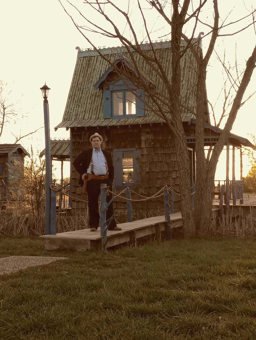 A cast member standing on the footbridge in front of the shingled cottage at sunset