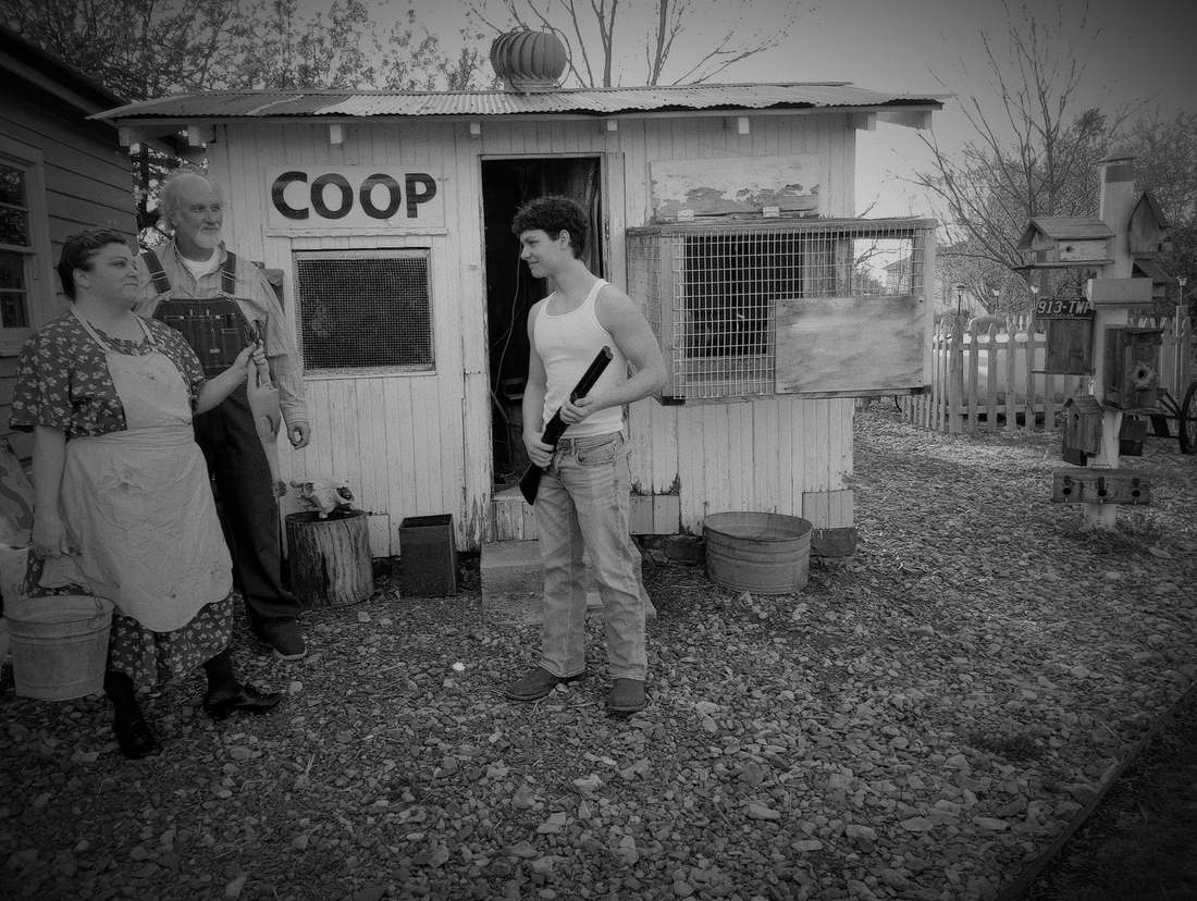 Three cast members outside a chicken coop, one holding a shotgun (black and white)