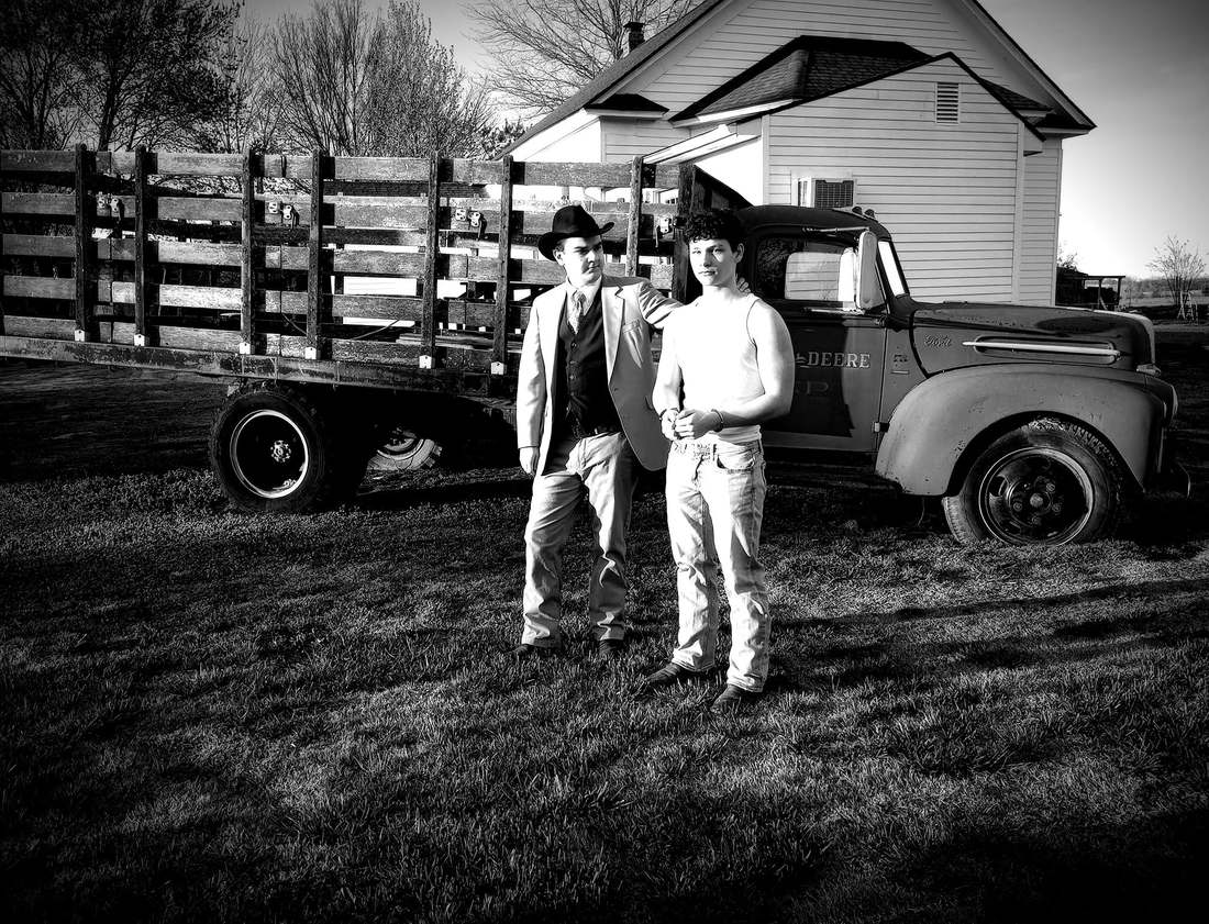 Two cast members standing in front of a vintage stake-bed farm truck (black and white)
