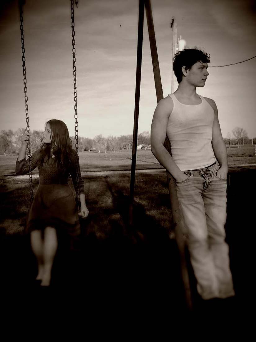 A cast member on a swing while another leans against the swing set frame at dusk (sepia)