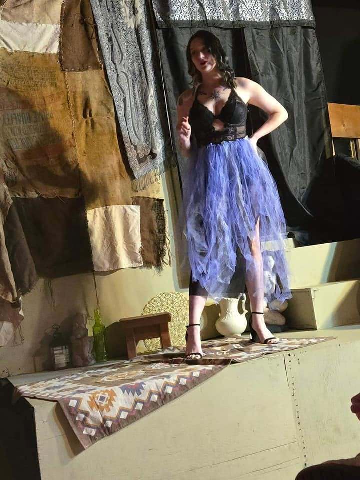 The storyteller poses atop the platform in a black top and layered blue skirt in Twisted