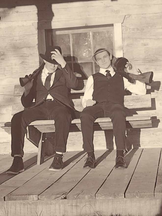 Two cast members with Tommy guns sitting on a cabin porch bench (sepia)