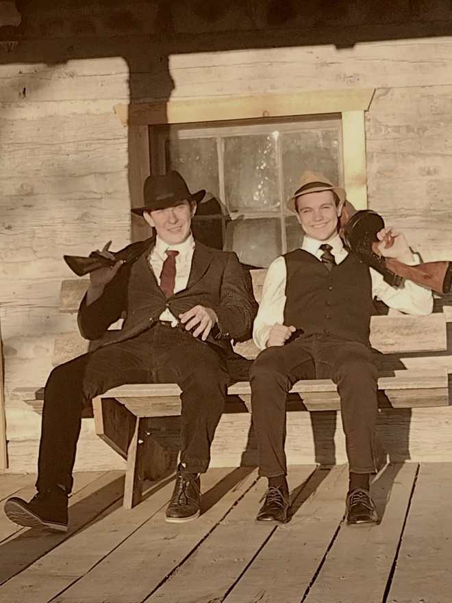 Two cast members with Tommy guns smiling on a cabin porch bench (sepia)