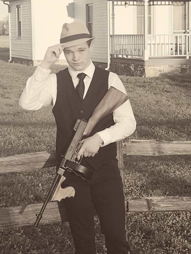 Clyde tipping his fedora and holding a Tommy gun by a split-rail fence (sepia)