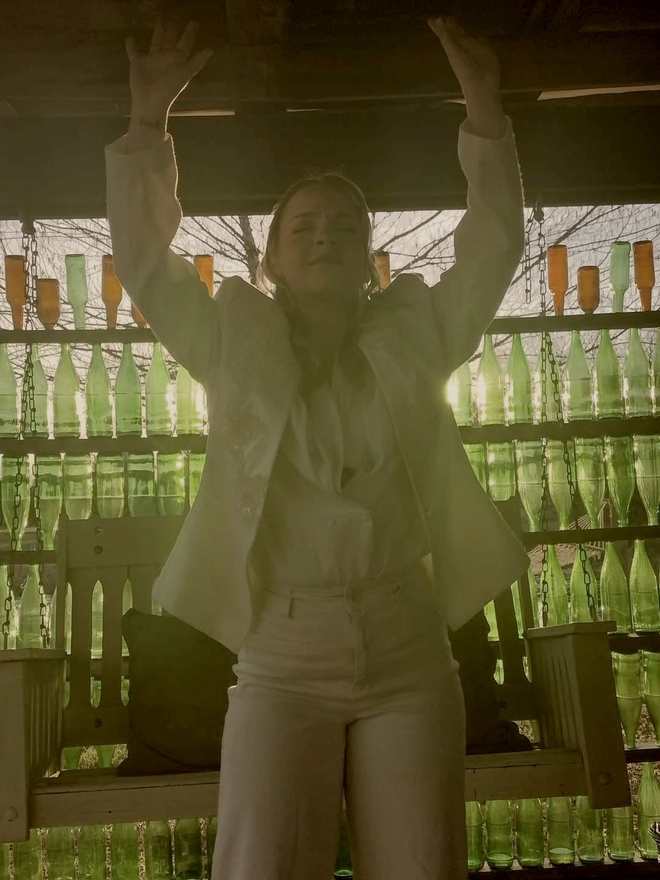 A cast member in a white suit backlit by the green bottle wall, arms raised