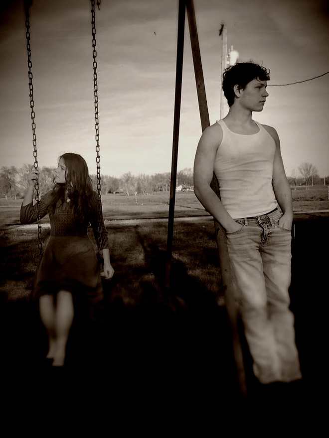 A cast member on a swing while another leans against the swing set frame at dusk (sepia)