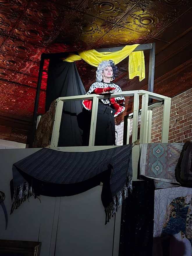 A grey-wigged performer in ruffles watches from the balcony railing in Twisted