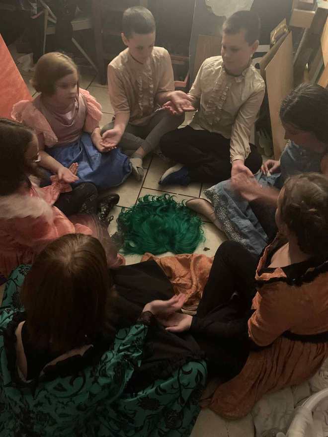 The cast huddles in a backstage circle around the creature's green wig on the floor