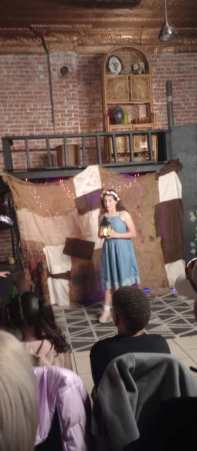 A young performer in a blue dress holds a lantern center stage before the patchwork backdrop