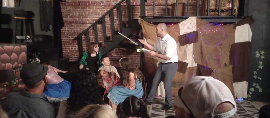 A performer gestures over an open book, telling the tale to the seated young cast