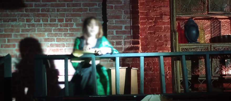 A performer reads from a book on the upper balcony, lit against the brick wall