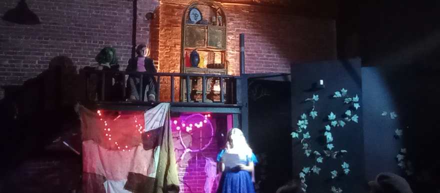 Two figures watch from the shadowy balcony as the scene plays below