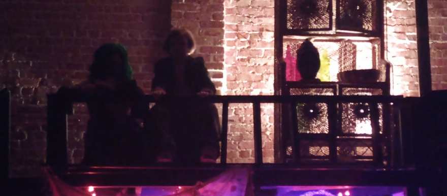 Two performers sit silhouetted on the balcony against the warm-lit brick wall