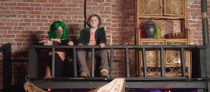 The creature, in a green wig and makeup, reads beside a young performer on the balcony