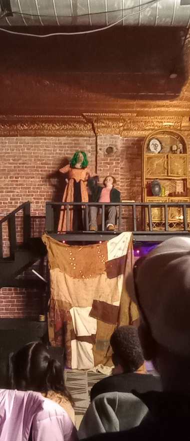 The creature stands on the balcony beside a seated performer, the patchwork curtain below