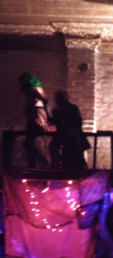 Two figures on the balcony in near-darkness above the pink-lit curtain