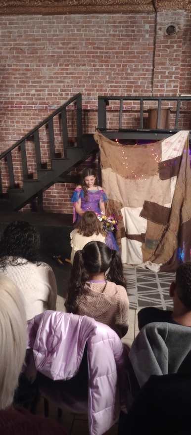 A performer in a purple dress holds a bouquet before the patchwork backdrop
