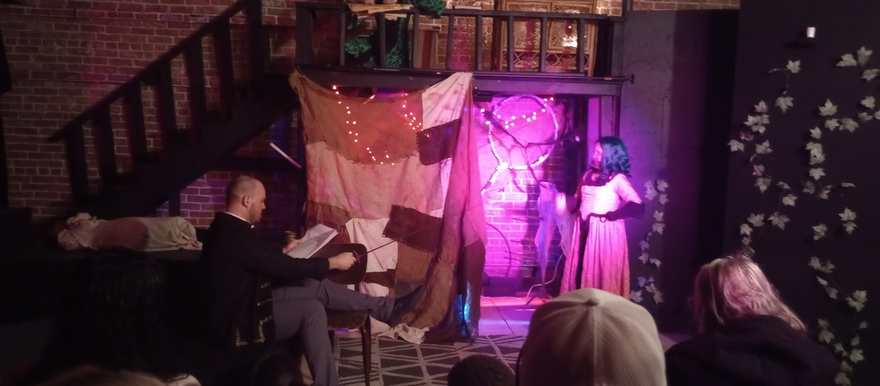 A performer in a long pink dress stands in the glowing pink doorway while another reads nearby