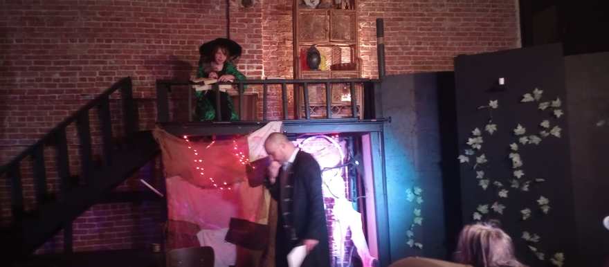 A performer in a wide-brimmed hat reads from the balcony as a scene unfolds below