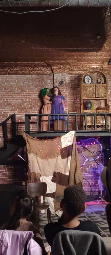 Two performers stand together on the balcony above the patchwork curtain
