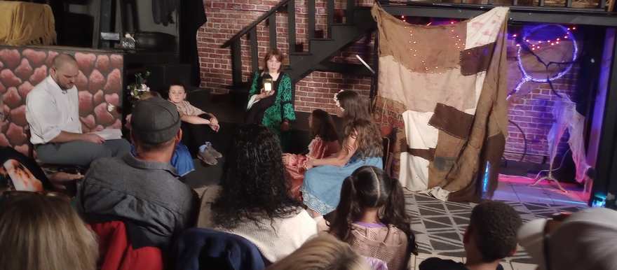 The company gathers on stage around the storyteller in green