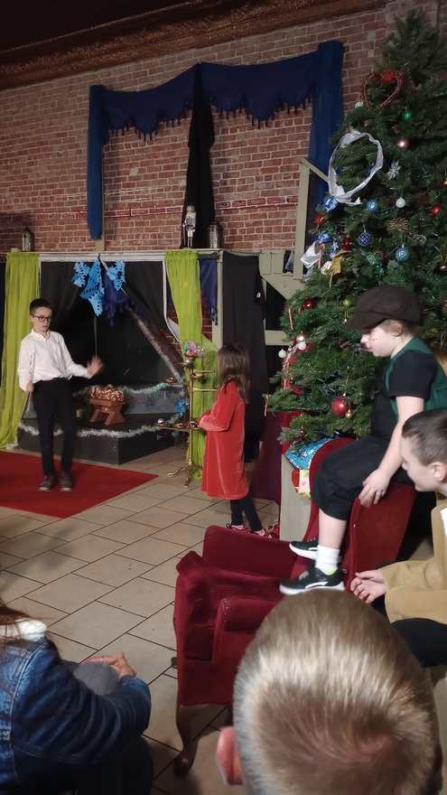 Young performers in costume gather beside the Christmas tree — a girl in red, a boy in a white shirt by the set