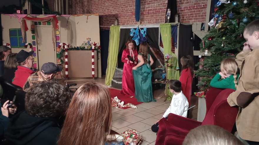 A wide view of the party scene, the gowned performers center stage and a young cameraman at the edge of the frame