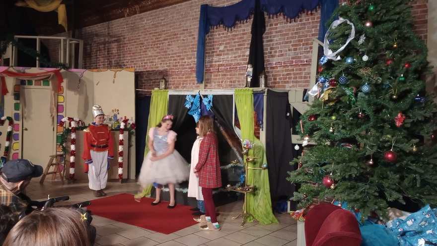 Clara in her white dress stands with the Nutcracker and a castmate in red pajamas beside the Christmas tree