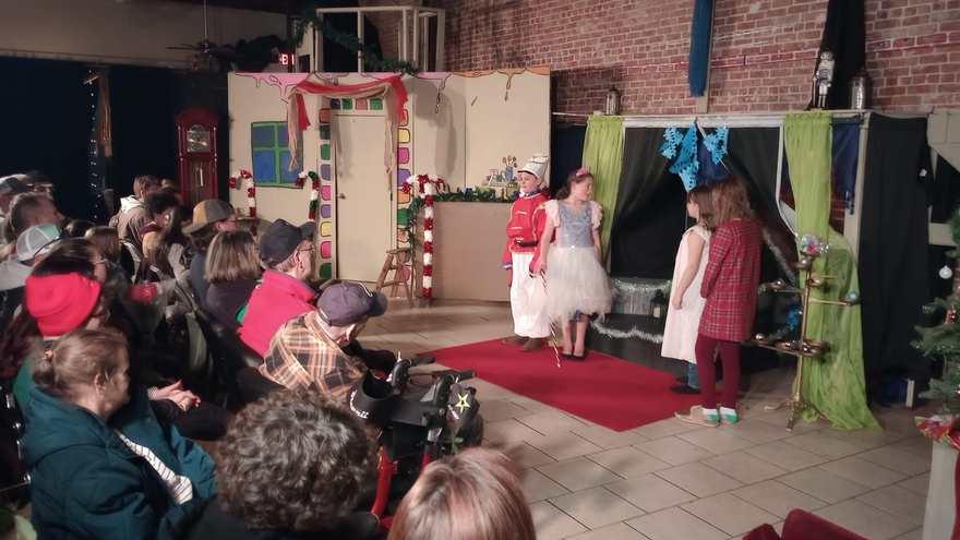 Performers in white and red play a scene on the red carpet as the audience watches from all sides