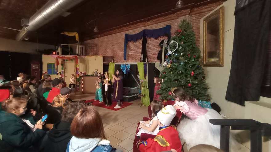 Performers stand center stage in a side view from the house, the seated cast watching from the sofa