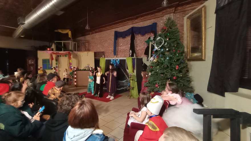 The cast in costume lounge on the sofa at the side of the stage, watching the scene across the room