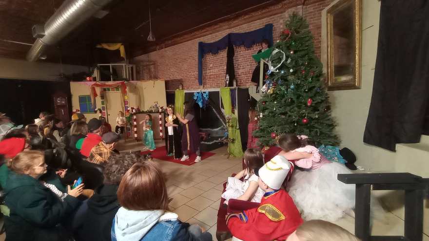 A wider side view — performers at the far set, the costumed cast watching from the sofa