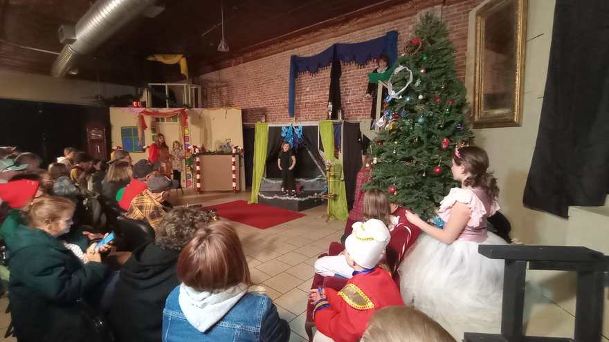 A young performer in a pink and white tutu dress sits on the arm of the sofa, watching the scene