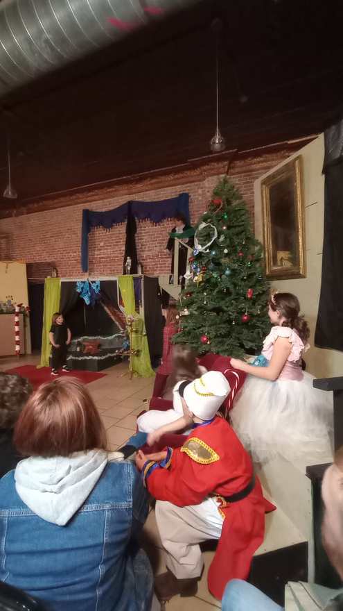 The Nutcracker sits beside Clara in her white dress by the Christmas tree as the audience watches