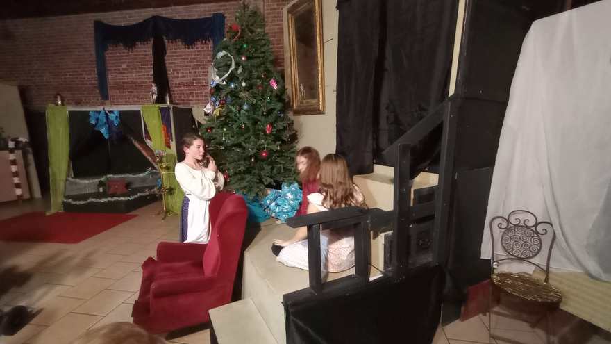 Behind the set, a young performer in a white nightgown waits by the red armchair