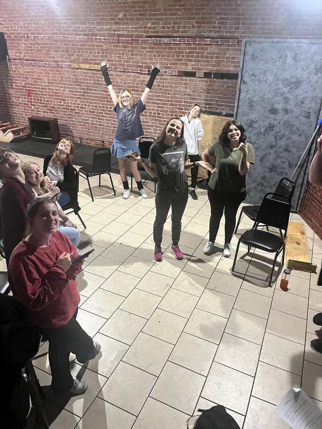 A Firebringer performer leaps with both arms raised in rehearsal as castmates watch in the brick-walled studio