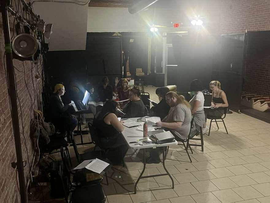 The Firebringer cast seated around tables in the darkened studio during rehearsal