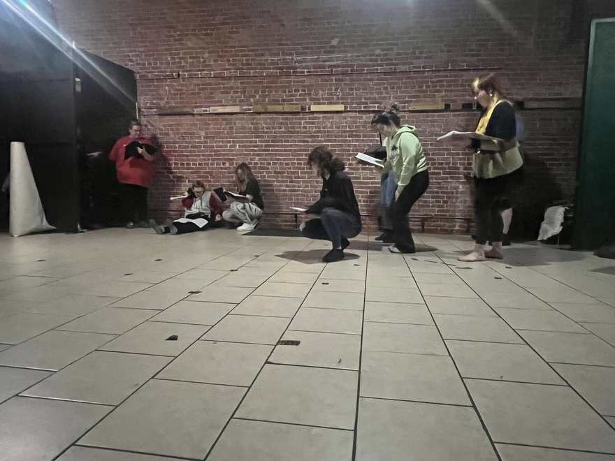 Firebringer performers crouch along the brick wall in rehearsal while two castmates stand with scripts